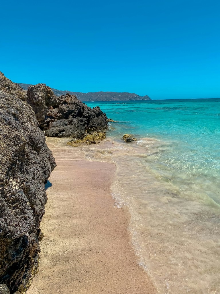 Pink-tinted sand and turquoise water at the rocky edge of Elafonissi Beach, Crete