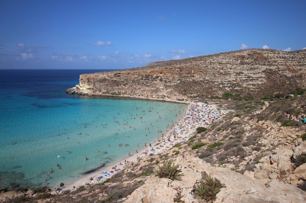 Crowded Rabbit Beach on Lampedusa island, Sicily, with turquoise water and rocky hillside